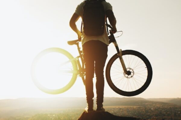 man-holding-mountain-bike-backlit-by-sunlight-standing-at-the-top-of-a-trail